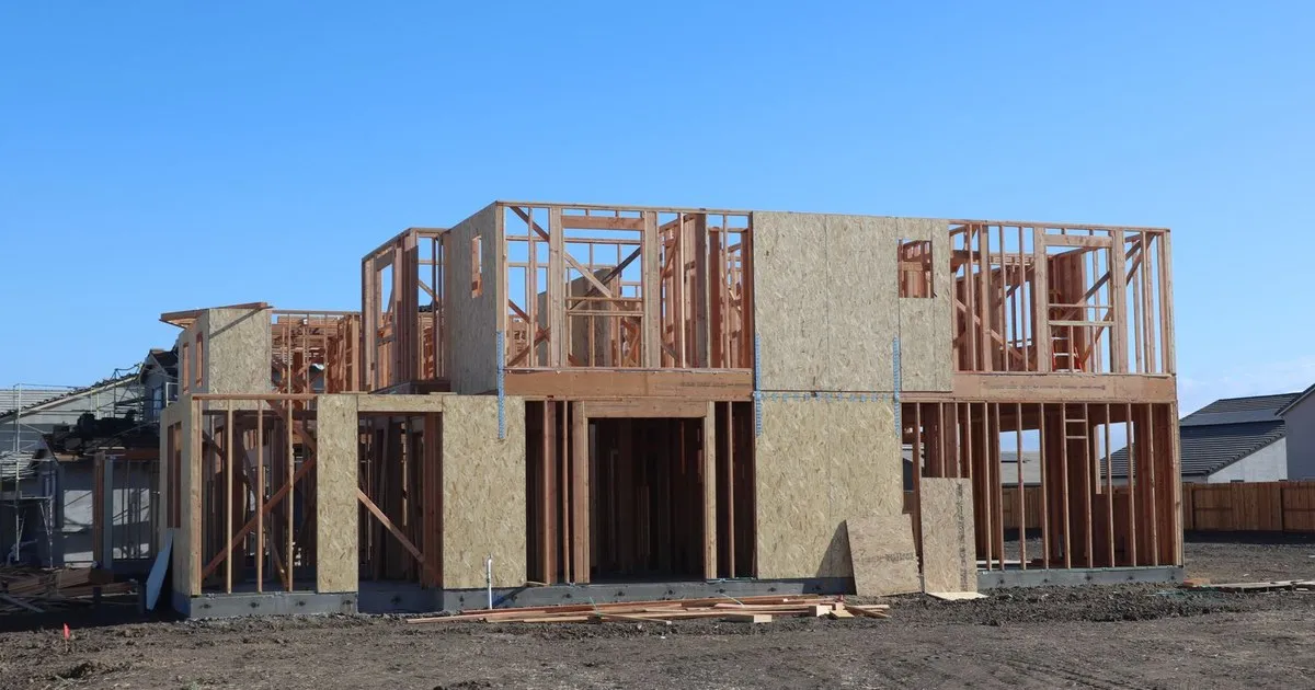 Wood-framed two-story house under construction on a cleared dirt lot under a clear blue sky