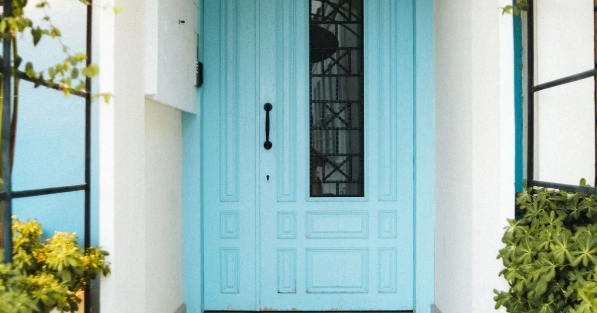 Light blue front door of a white house with green plants on each side
