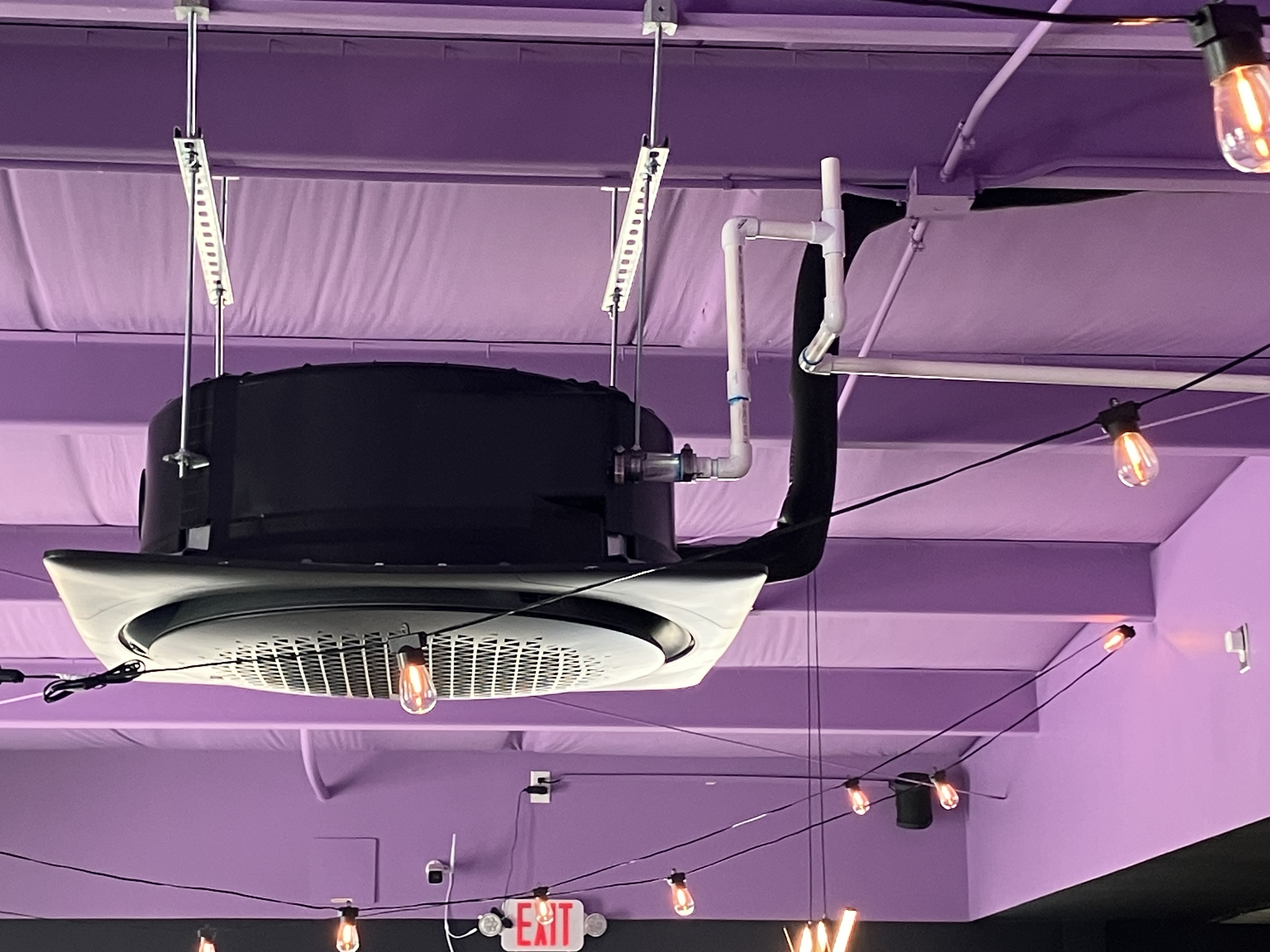 Ceiling-mounted air conditioning unit with black casing and visible ductwork in a purple-themed interior space.