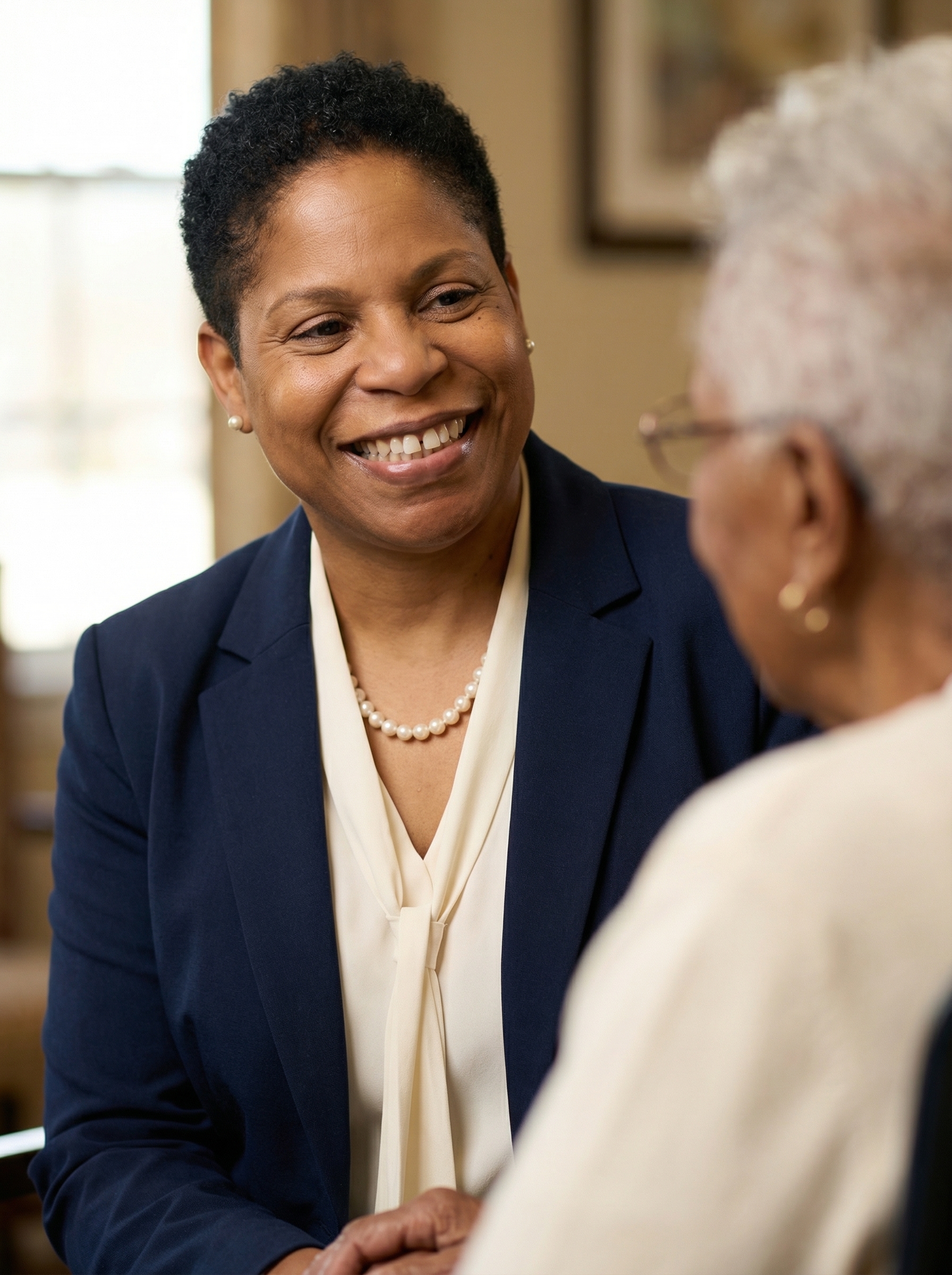 Elderly woman and adult daughter, both smiling, sitting together on a beige sofa with leafy green plants in the background, natural light.