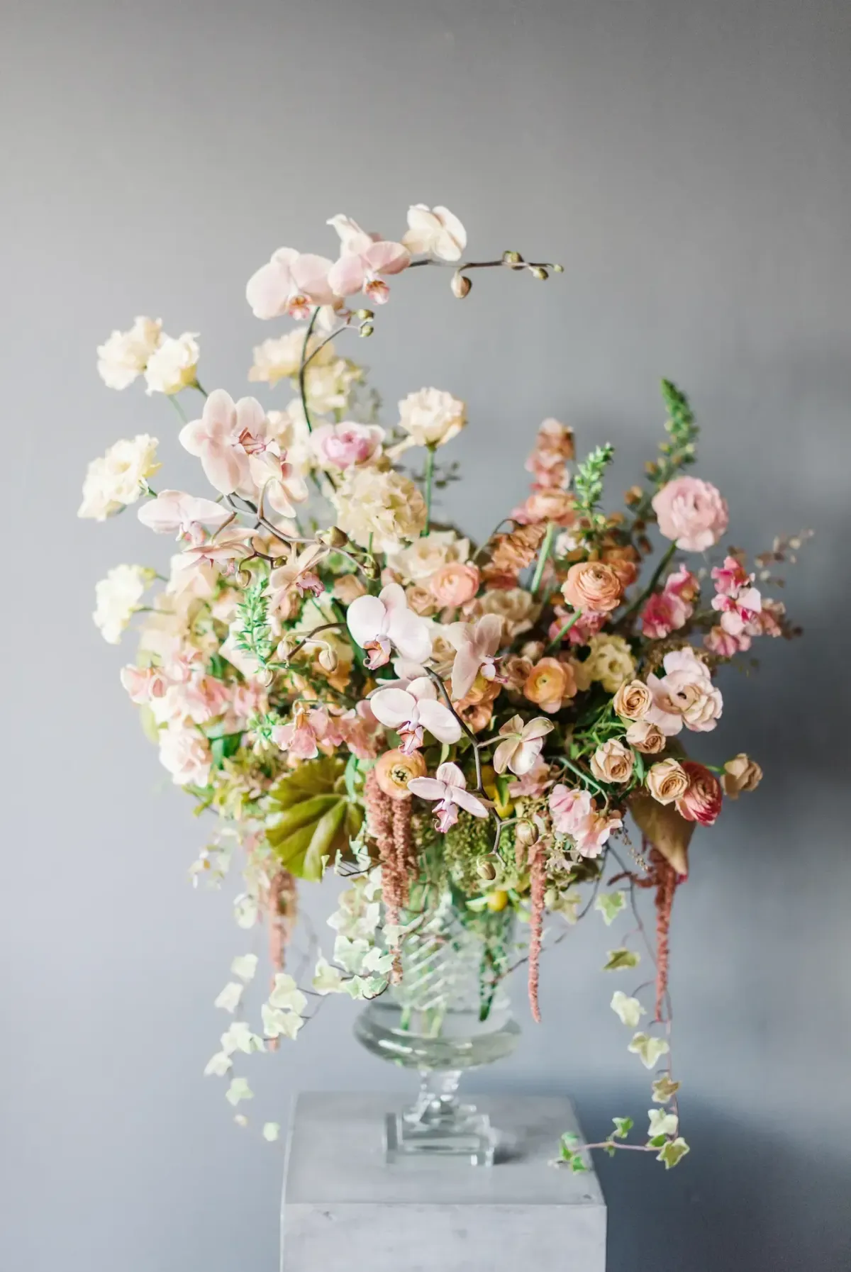 A glass vase filled with an arrangement of pastel flowers, including roses and orchids, against a gray background.