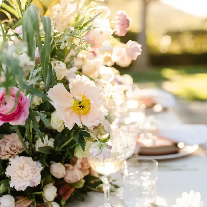 A bright floral centerpiece with pink, yellow, and white flowers on an outdoor dining table set with glassware and plates in soft sunlight.