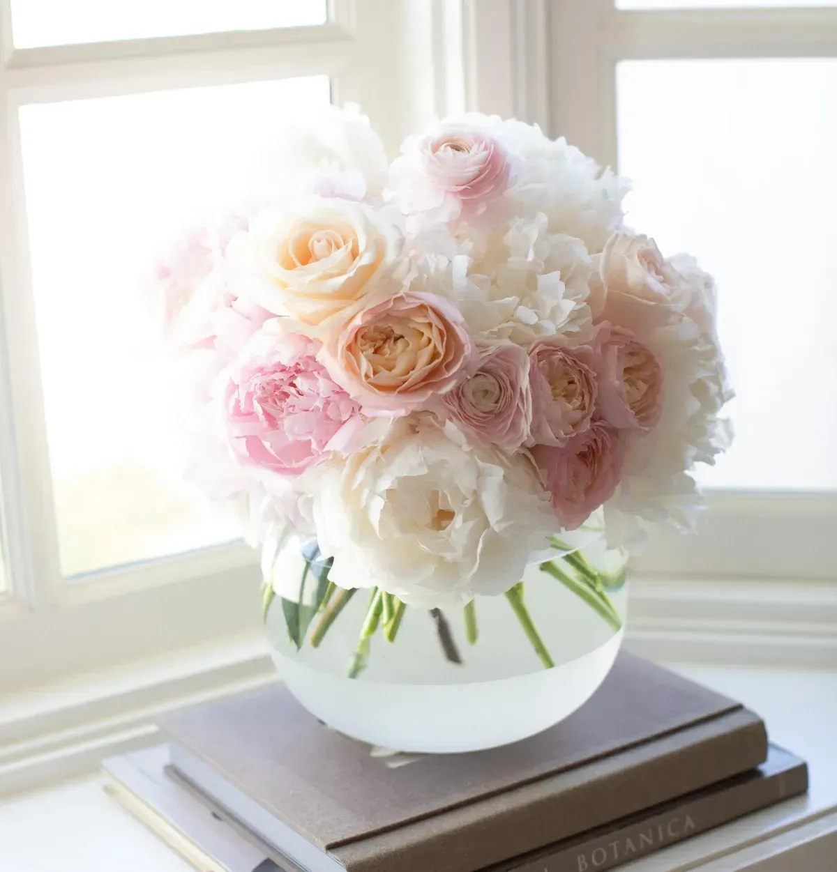 A round glass vase filled with soft pink and white roses, placed on stacked books by a bright window.
