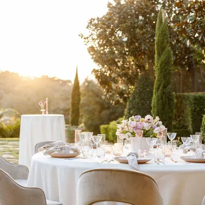 Elegant outdoor dining table set with glassware, white plates, and a centerpiece of pink flowers, with tall greenery and a sunset in the background.
