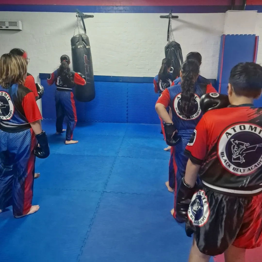 Junior students wearing Atomic uniforms and boxing gloves practise on hanging punch bags in the blue-matted training room at Atomic Black Belt Academy in Coventry.
