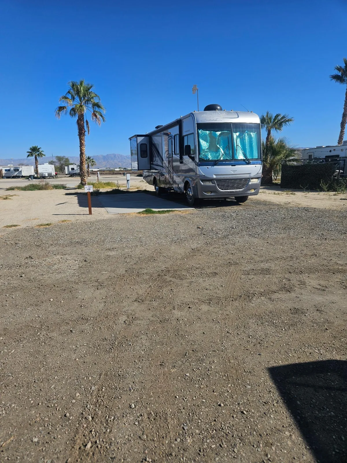 A white RV is parked on a dirt lot under a clear blue sky. Other RVs are visible in the background, along with palm trees.