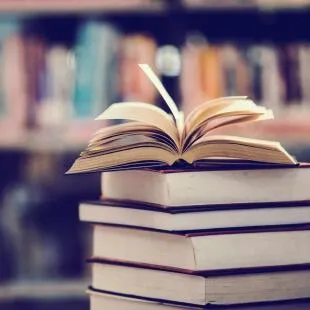 A stack of books sits on a table with an open book on top, against a blurred background of bookshelves.