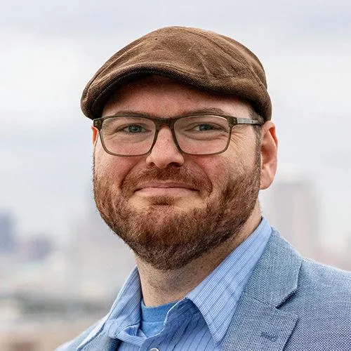 Portrait of Jesse Sieger-Walls wearing glasses, a cap, and a blazer.