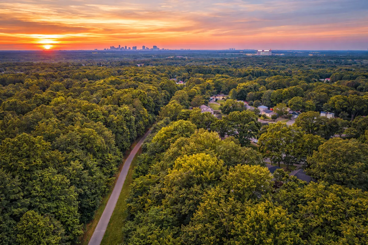 A vibrant, wide-angle aerial shot of Mableton, Georgia, showcasing the lush green tree canopy that the area is known for. The Silver Comet Trail is visible as a clean, winding path through the trees. In the distance, the Atlanta skyline and the Truist Park (The Battery) lights are visible on the horizon, emphasizing the city's close-in location. The sun is setting, casting a warm orange and purple glow over the suburban rooftops.