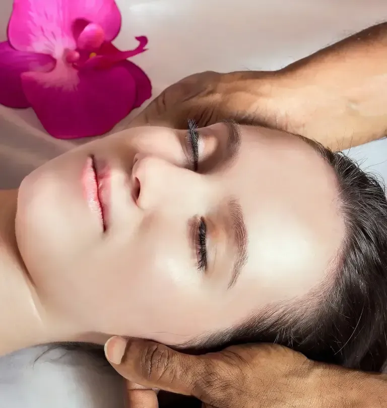 A person is enjoying a soothing facial massage while lying down with their eyes closed, a pink flower delicately placed near their head.