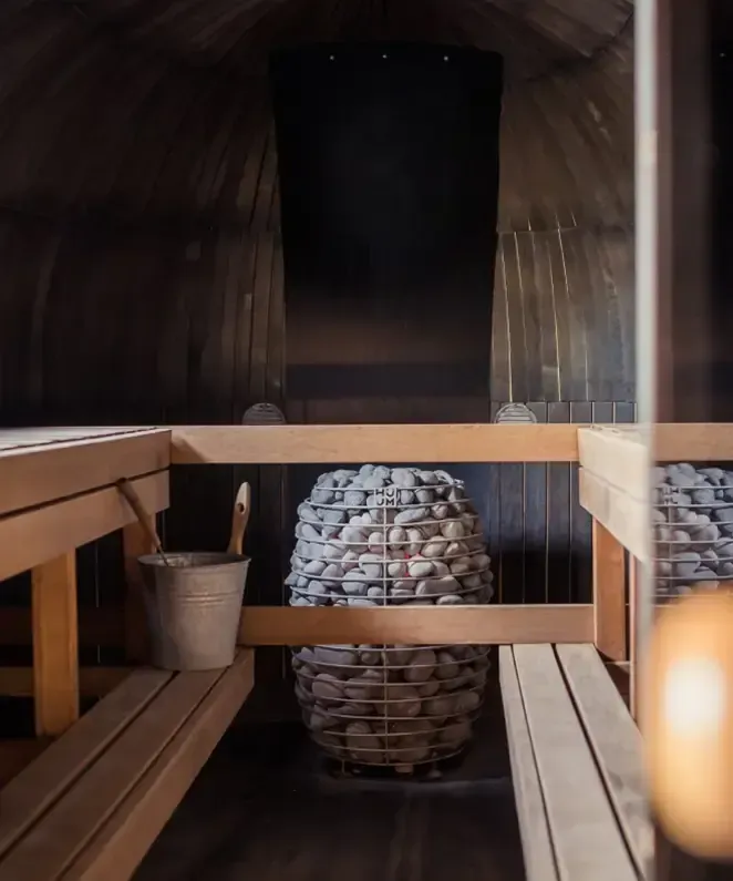 The interior of a traditional wooden sauna features wooden benches along the walls and a bucket beside a pile of sauna stones in the center, creating an atmosphere where massage essence aromas can enhance the soothing experience.