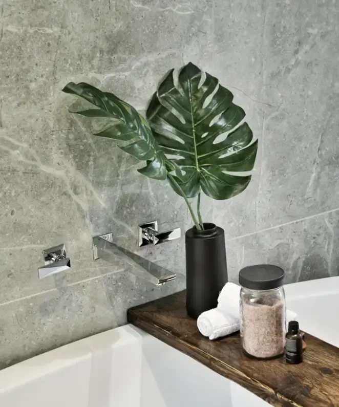 A modern bathroom features a wall-mounted faucet, a black vase with large green leaves, a jar of bath salts, a rolled white towel, and a small bottle on a wooden board over a white bathtub.