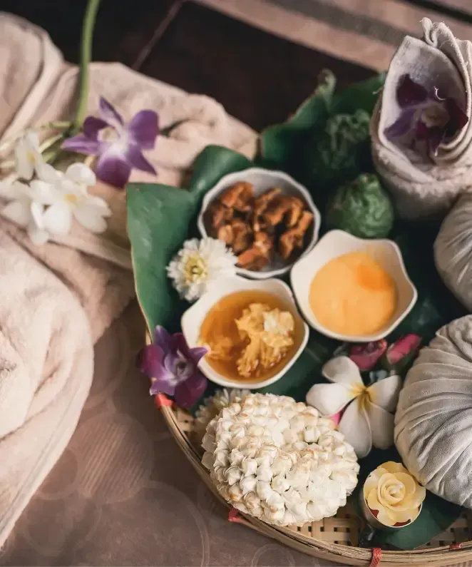 A spa setup featuring a towel, flowers, and a wooden tray with various spa items: bowls of honey and a yellow substance, dried fruits, herbal spheres, and flower decorations.