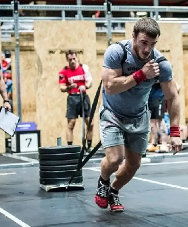 A man in athletic wear pulls a heavy sled with weights in a gym, while another person in the background observes and takes notes.