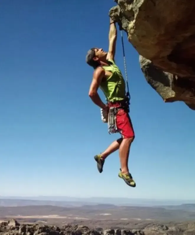 A rock climber in red shorts and a green tank top hangs by one hand from a rock ledge, with a safety rope attached. Against the backdrop of the desert landscape and clear blue sky, he dreams of offering massage services for mountain climbers &amp; hikers after such an exhilarating ascent.