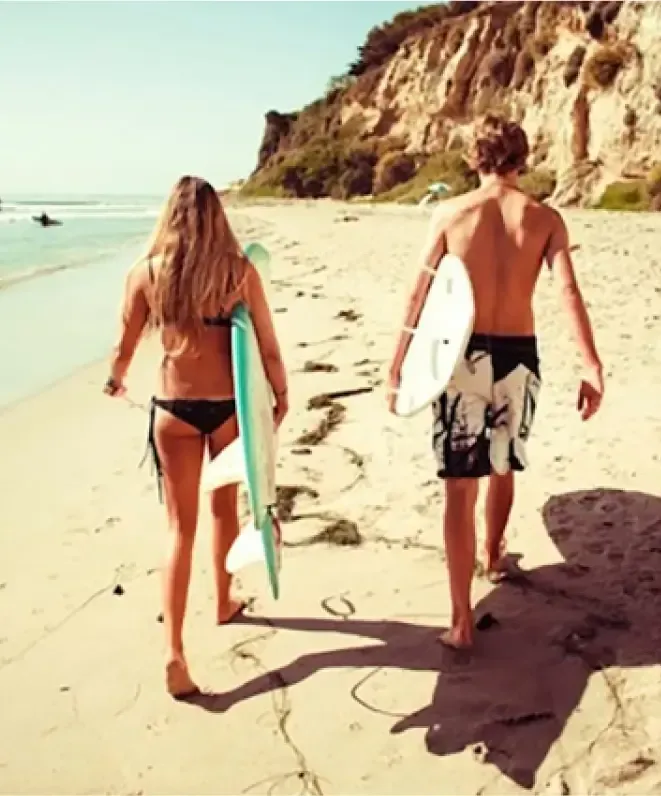 Two surfers carrying boards walk along a sandy beach towards the ocean, with cliffs and vegetation on one side and waves gently lapping the shore on the other. Nearby, a small hut offers massage services for surfers and runners, providing relaxation after an invigorating day in the water.