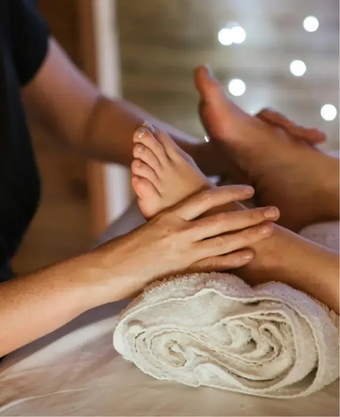 Close-up of a person receiving a foot massage, with hands gently massaging the foot. The setting is dimly lit, enhanced by the soothing scent of massage essence and highlighted with a soft focus background and small, decorative lights.