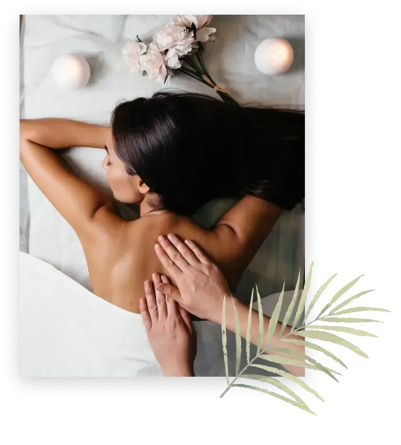 A person lying face down on a massage table, receiving a back massage. The setting includes candles, flowers, and a palm leaf decor.A person lying face down on a massage table, receiving a back massage. The setting includes candles, flowers, and a palm leaf decor.