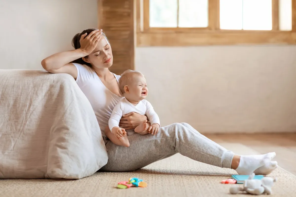 Exhausted mother sitting on floor holding crying newborn showing signs of postpartum depression — Medivira Frisco TX
