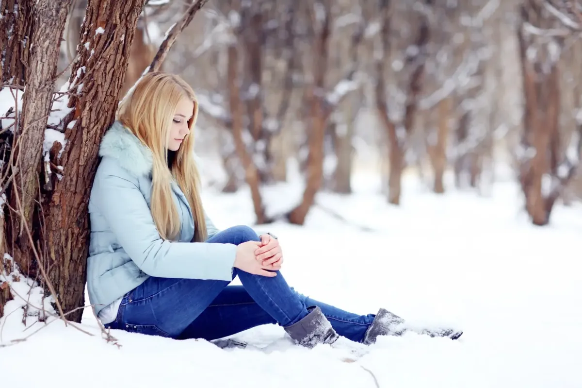 A young woman sitting alone in the snow, leaning against a tree in a bare winter forest, with her head bowed and hands clasped around her knees, conveying feelings of isolation and sadness associated with Seasonal Affective Disorder.