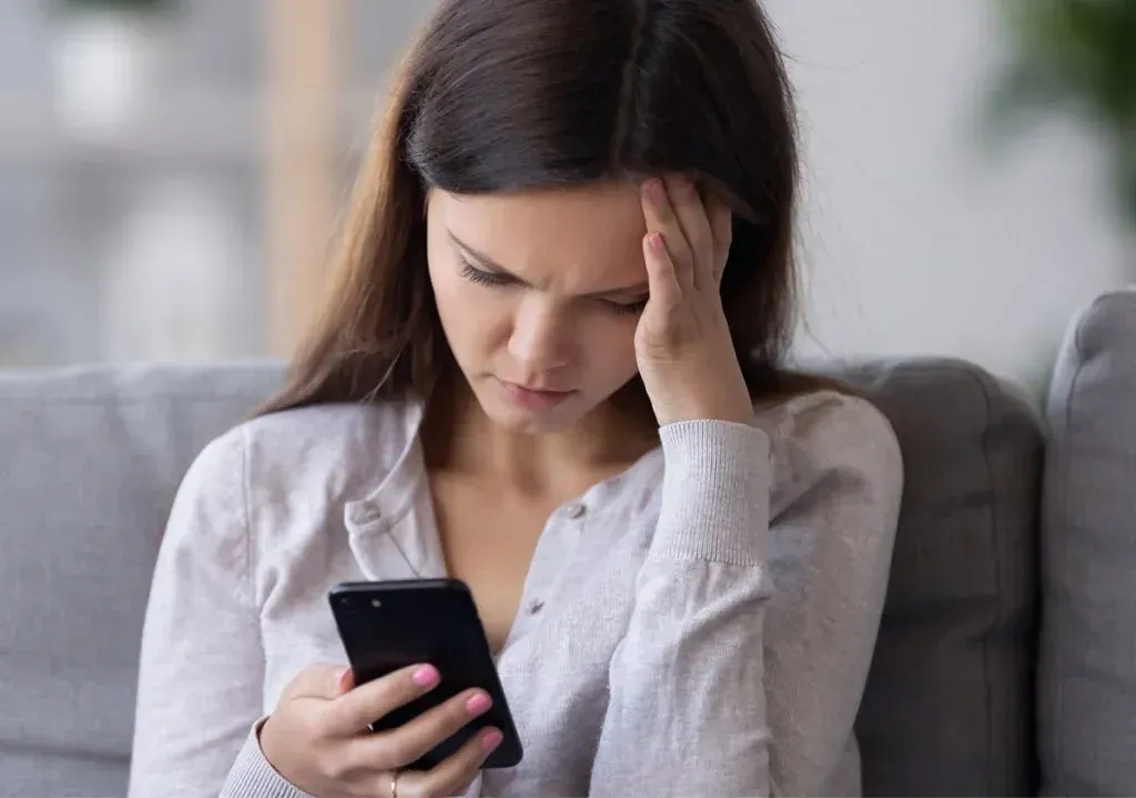 Stressed woman sitting on a couch holding her head while looking at her phone, representing emotional distress and the mental health impact of kratom addiction — Medivira