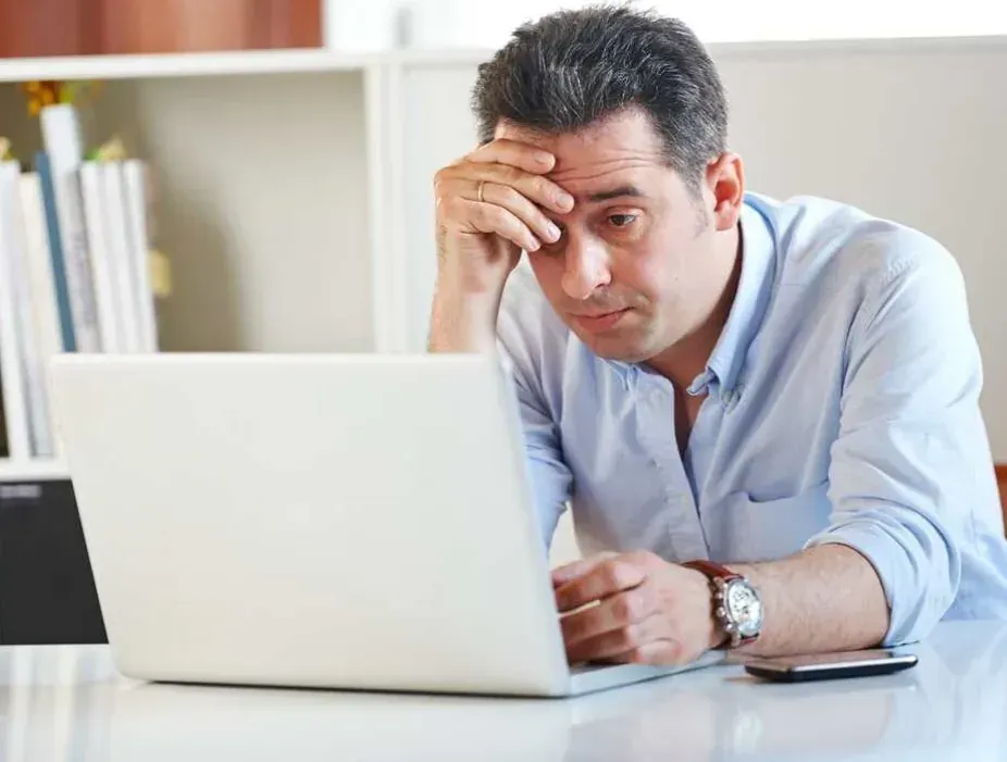 Stressed man sitting at a desk holding his head while staring at a laptop, illustrating the compulsive behavior and mental strain associated with internet addiction — Medivira