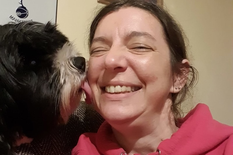 A smiling woman in her 30s, holding a notepad, with a border collie at her side, standing in a sunlit home office. The setting is cozy and professional, reflecting her role as a canine behaviourist.
