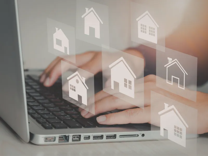 Hands typing on a laptop with house icons floating above the keyboard, representing an online home search
