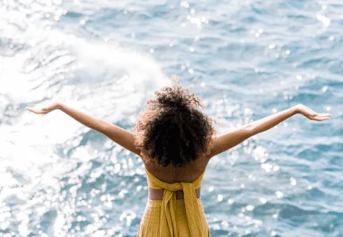 Woman of color in yellow dress facing ocean with out stretched  arms