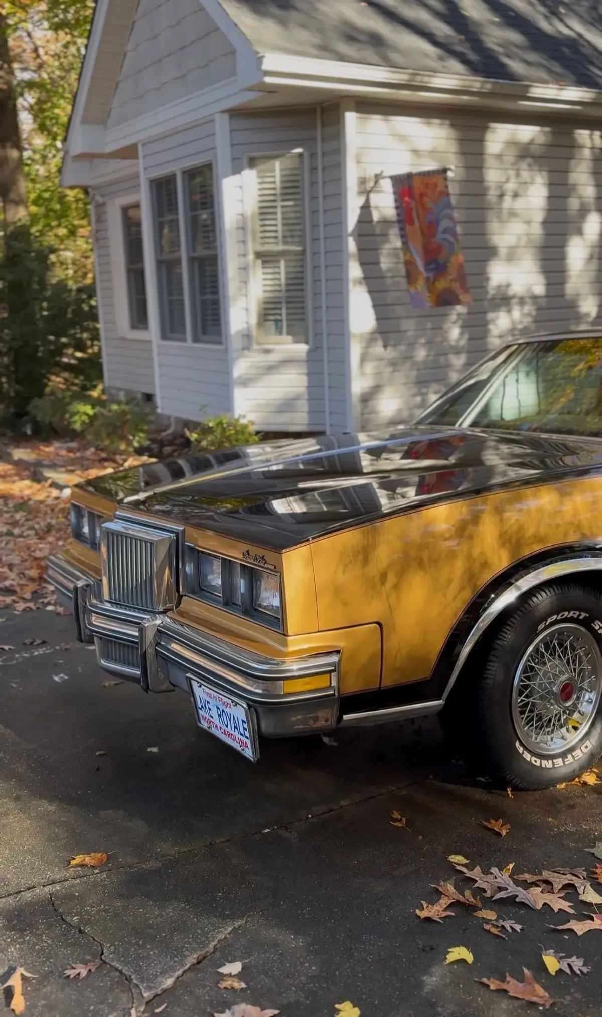 Golden and black classic car parked in front of a white house with a fall-colored flag.