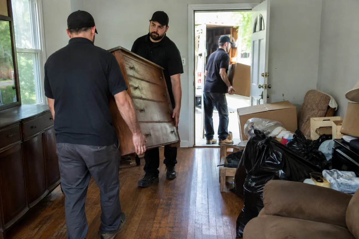 Property cleanout crew carrying an old dresser and boxed belongings out of an estate-transition home.
