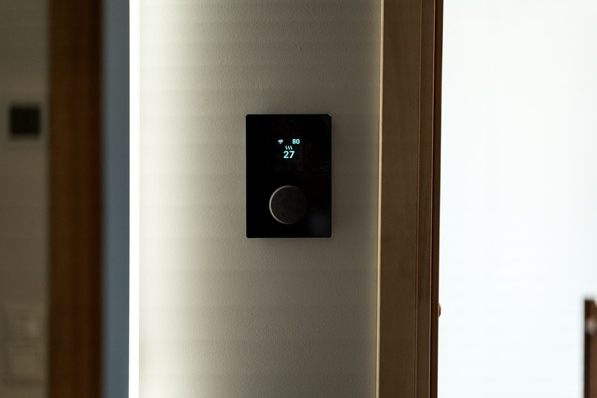 Modern black smart thermostat mounted on a wall in a home hallway, displaying WiFi status and temperature
