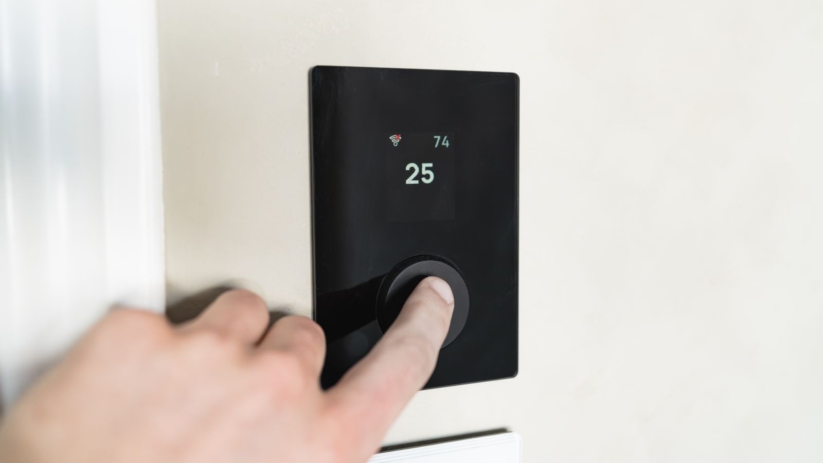 Finger pressing the dial on a black smart thermostat mounted on a light interior wall