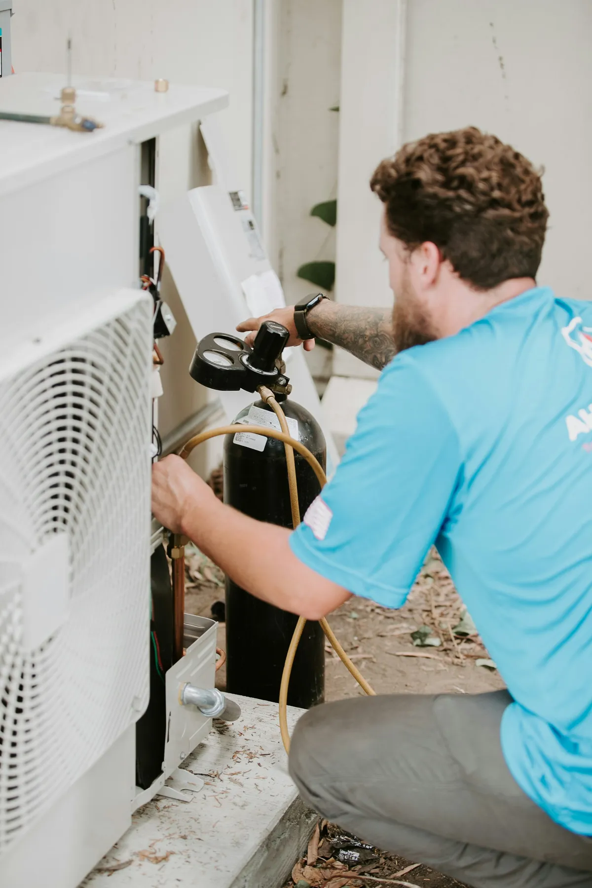 HVAC technician in blue shirt connecting a pressure gauge manifold to an outdoor AC condenser unit to check refrigerant charge and diagnose a leak