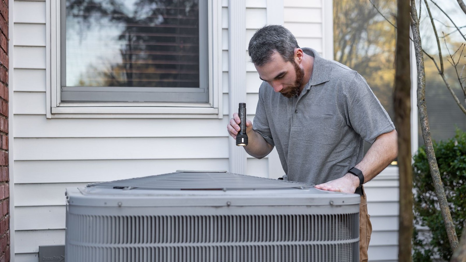 HVAC technician inspecting a residential outdoor AC condenser unit with a flashlight