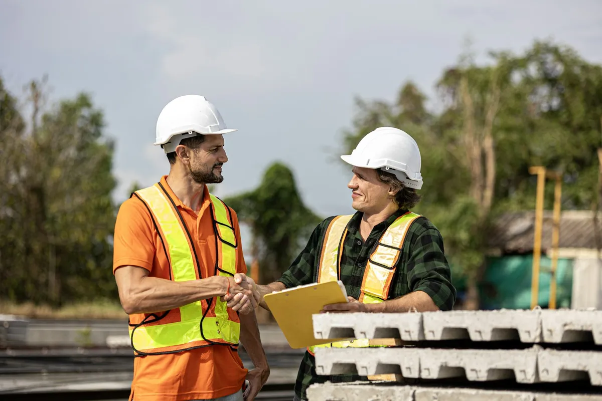 Two contractors in hard hats shaking hands over a signed quote at a job site