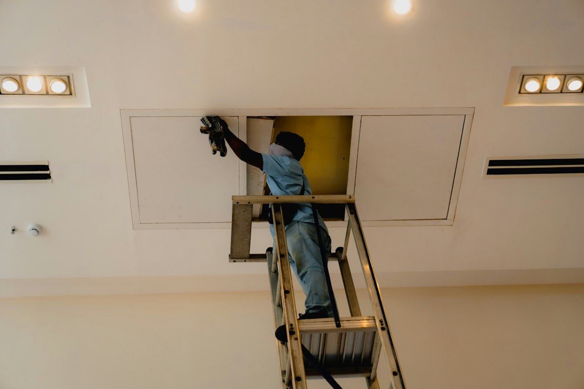 HVAC technician on a ladder inspecting a home's attic access panel for storm moisture damage