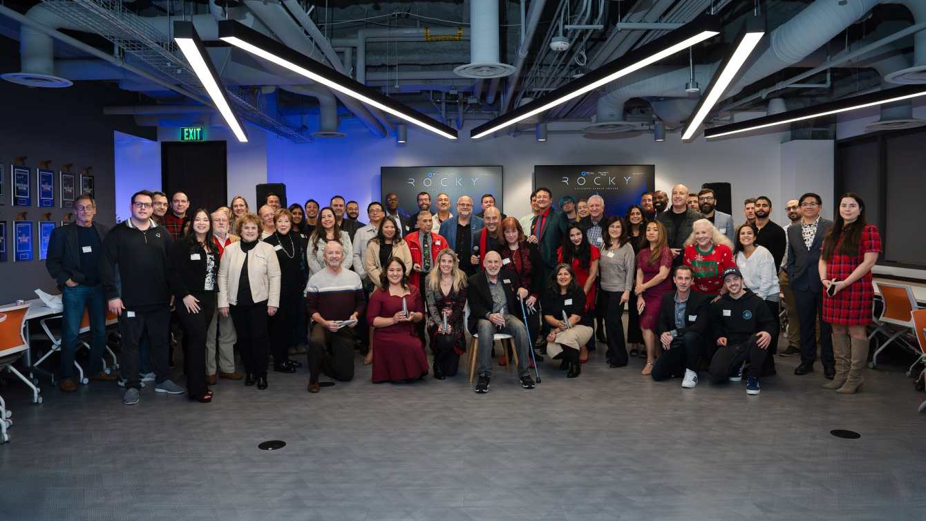 Group photo of TCVN founders, members, and award recipients at the Rocky Founders Choice Awards holiday celebration, with Rocky branding displayed on screens in the background