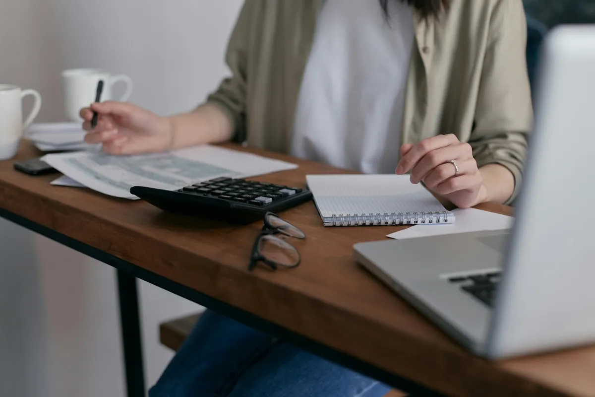 Person working at a desk with a laptop, calculator, notebook, and coffee mug, suggesting focused financial or administrative tasks.