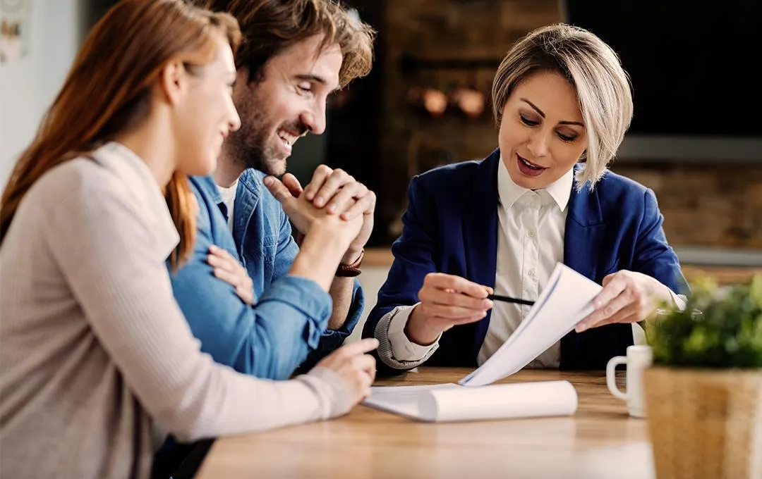 Professional woman in a suit explaining documents to a smiling couple across a table, indicating a business consultation or financial planning session.