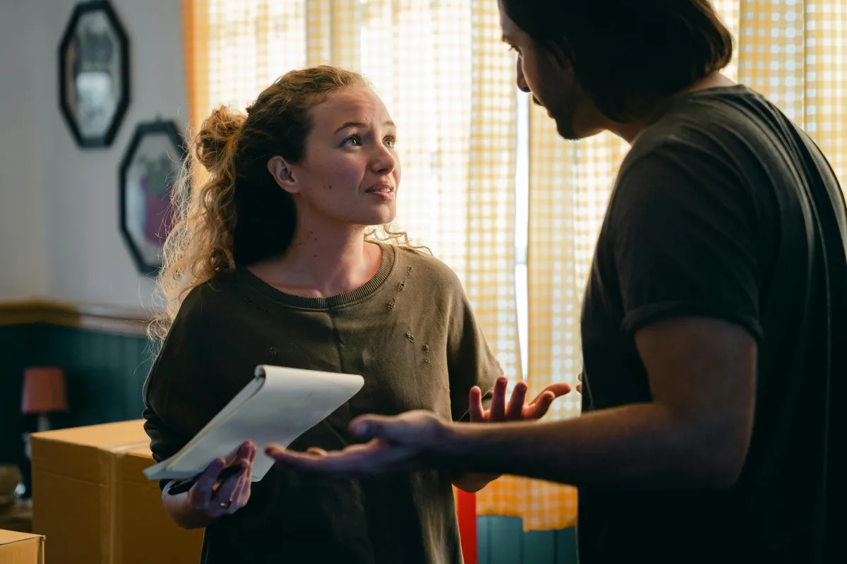 A couple inside a home surrounded by boxes, engaged in a serious discussion while one person holds a notepad, possibly planning or reviewing moving or financial details.