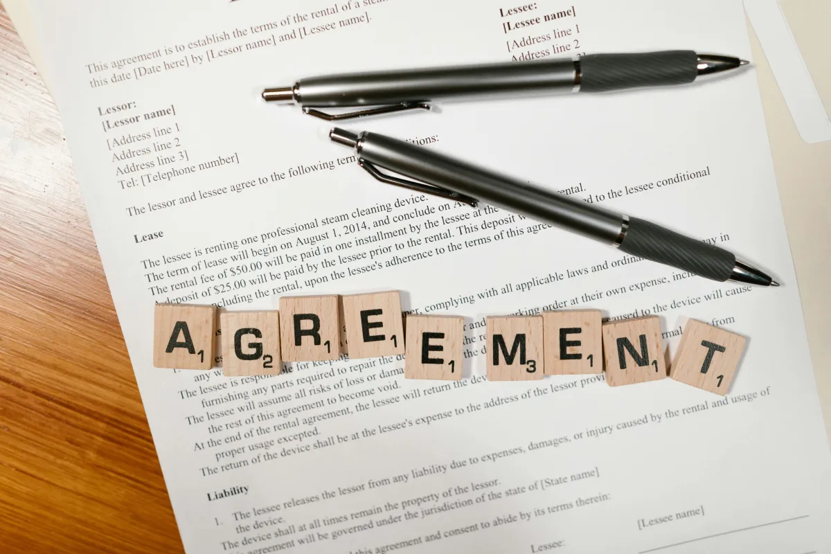 Close-up photo of a lease agreement document with two pens on top, and Scrabble tiles spelling the word "AGREEMENT" placed across the paper.