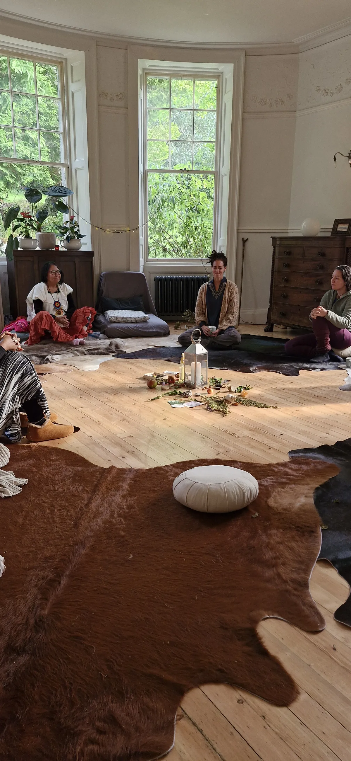 A woman sitting comfortably on the floor of a bright, sunlit room, engaged in an activity, with cushions, plants, and natural light.