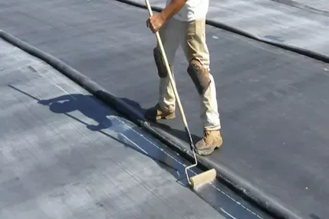Contractor rolling adhesive along a flat rubber roof seam during commercial waterproofing service