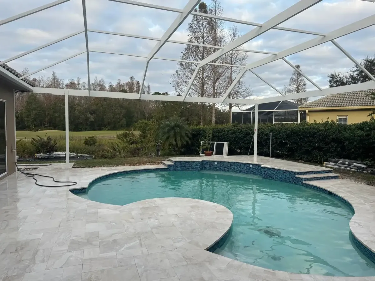 White metal frame of a pool enclosure installed above a tiled residential swimming pool with landscaping and trees in the background