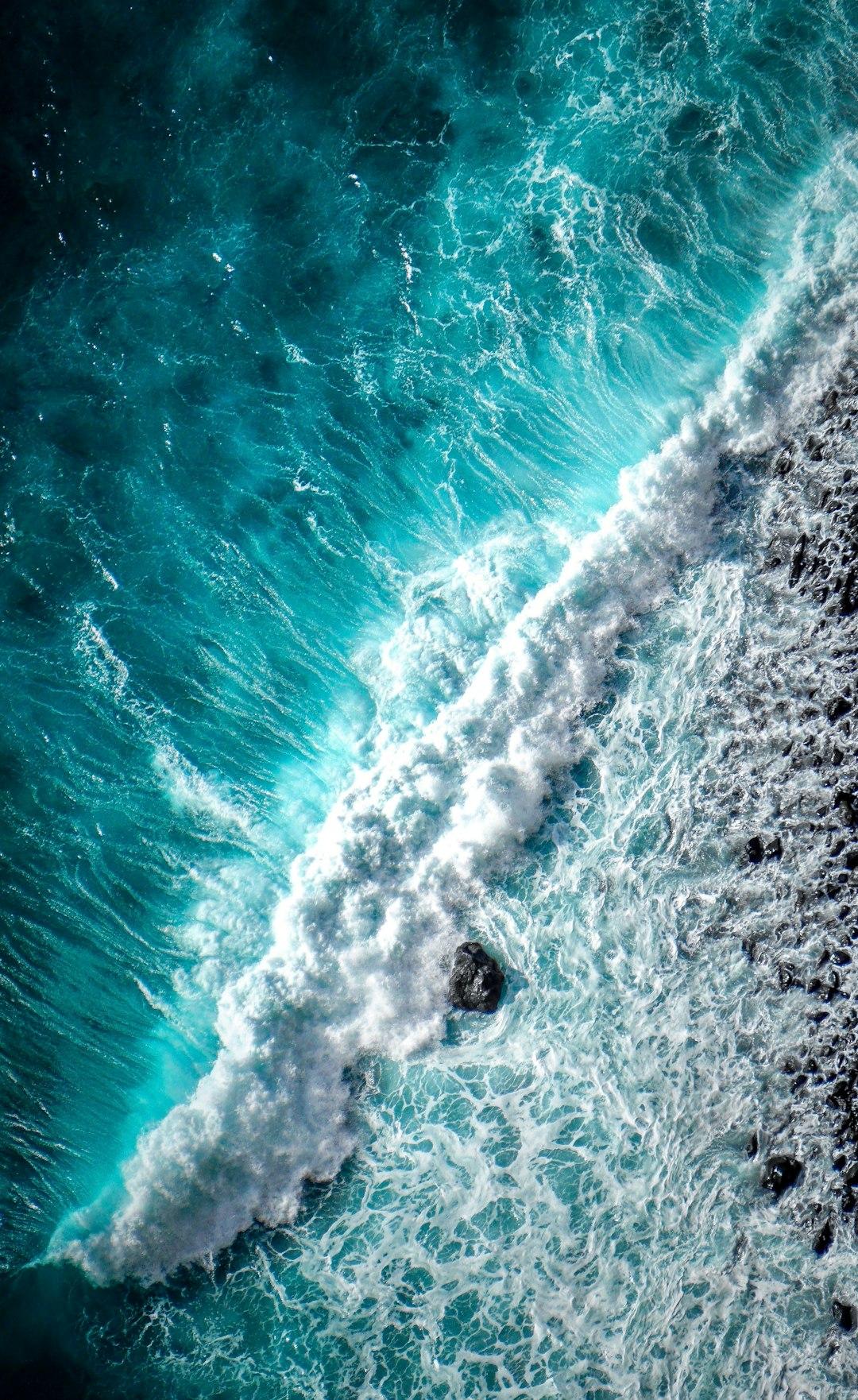 Waves crash toward the shore with hues of deep blue all the way to white in the wave. Black rocks present as the shoreline.