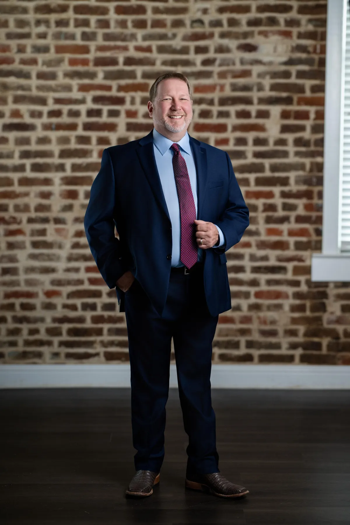 Professional middle-aged man in a navy blue suit and red tie standing confidently in front of an exposed brick wall, smiling for a business portrait.