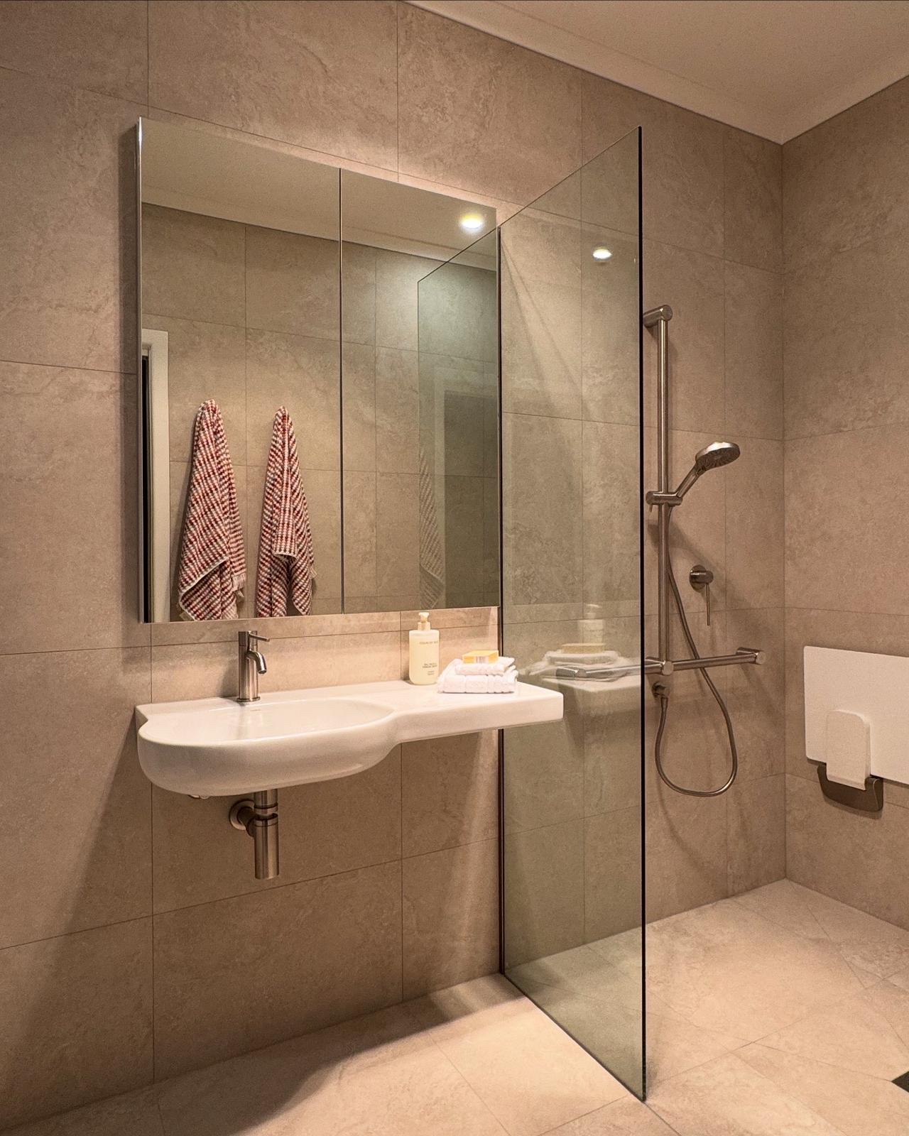 Generate an AI image of a wide-entry bathroom with a roll-in shower. Clean minimal design. Fold-down shower seat in a timber finish. Non-slip floor tiles that look intentional and premium. Soft natural light.