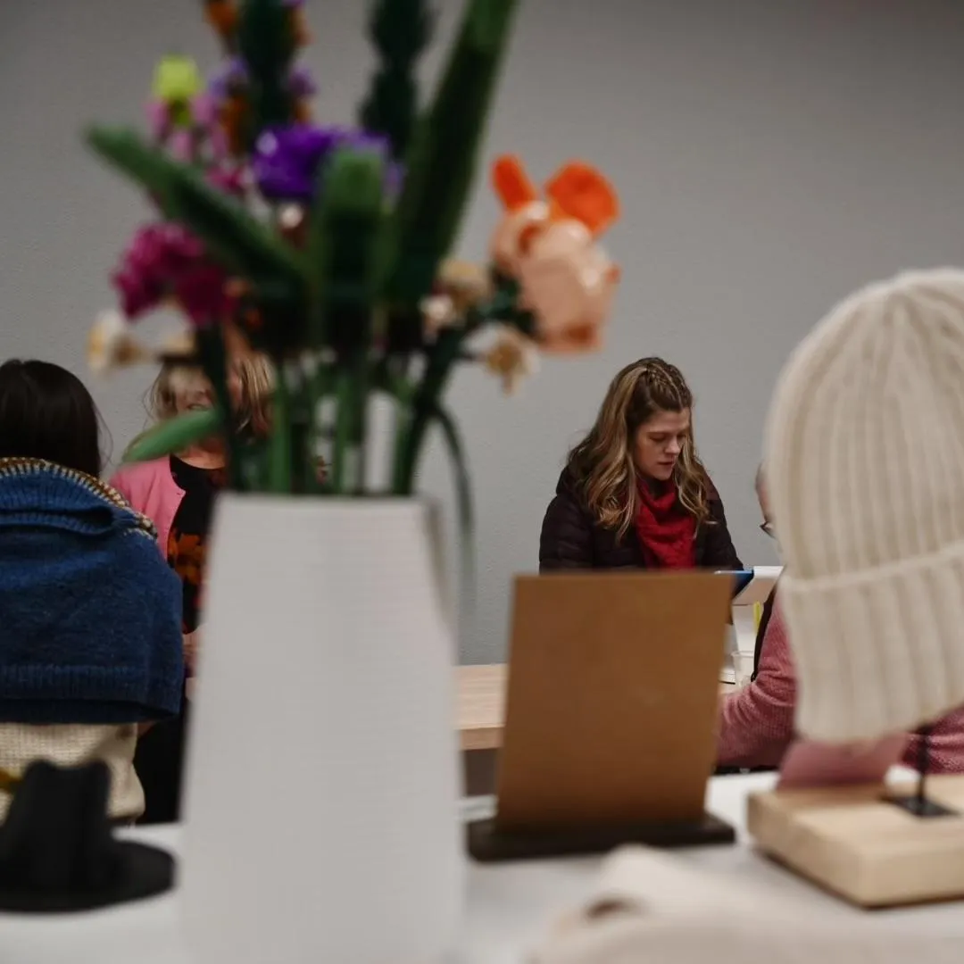 Woman with long hair in a red scarf engaged in conversation at a yarn shop, with colorful flowers and knitted items in the foreground, illustrating community and creativity in a local business setting.