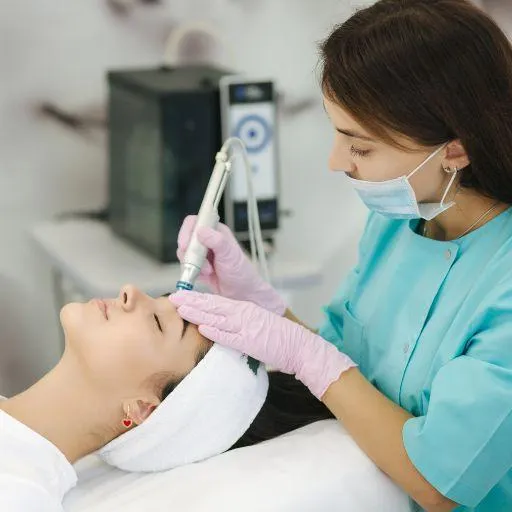 A professional performing a hydrafacial treatment on a person lying down with a towel on their head in a clinical setting.
